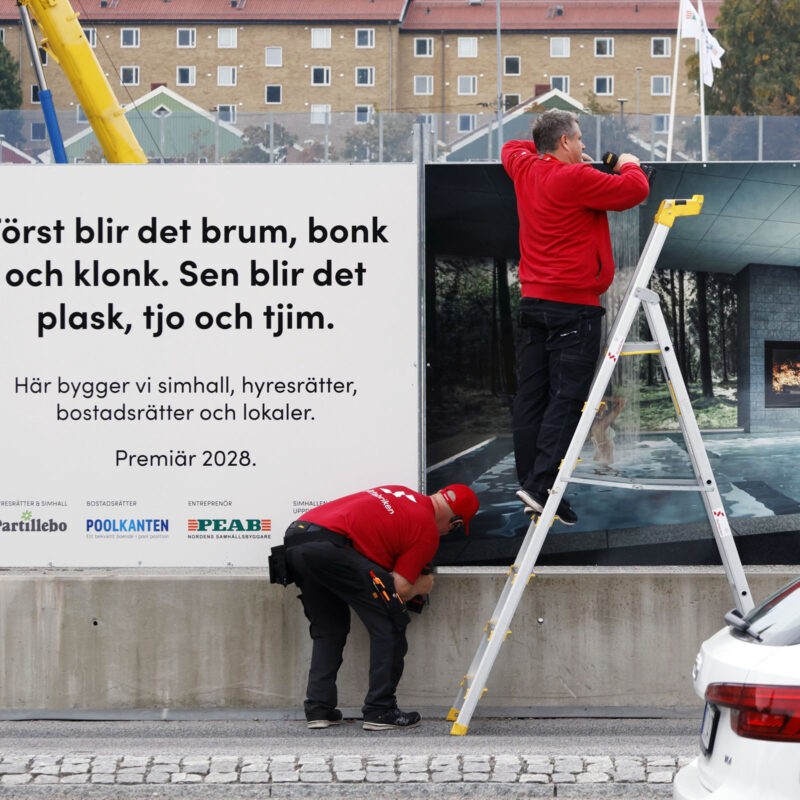 Två montörer med skyltfabriken profilkläder monterar en plåtskyltar mot ett byggstaket för Partille bo.