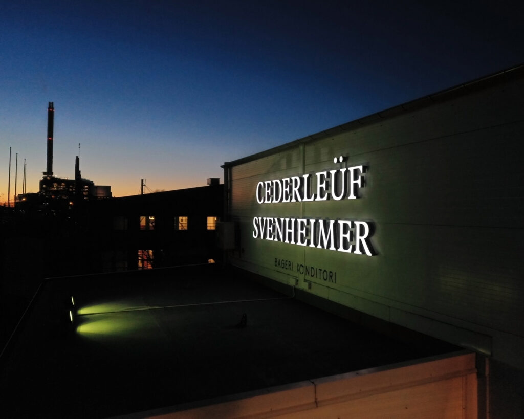 En nattbild över en fasadskylt som lyser vitt. På dagen är denna skylt svart.