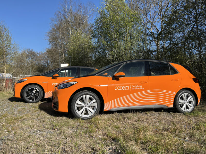 Två bilar står uppställda på en äng. Båda är helfolierade med en orange printad dekor med Corem fastigheter logotype på sidan.