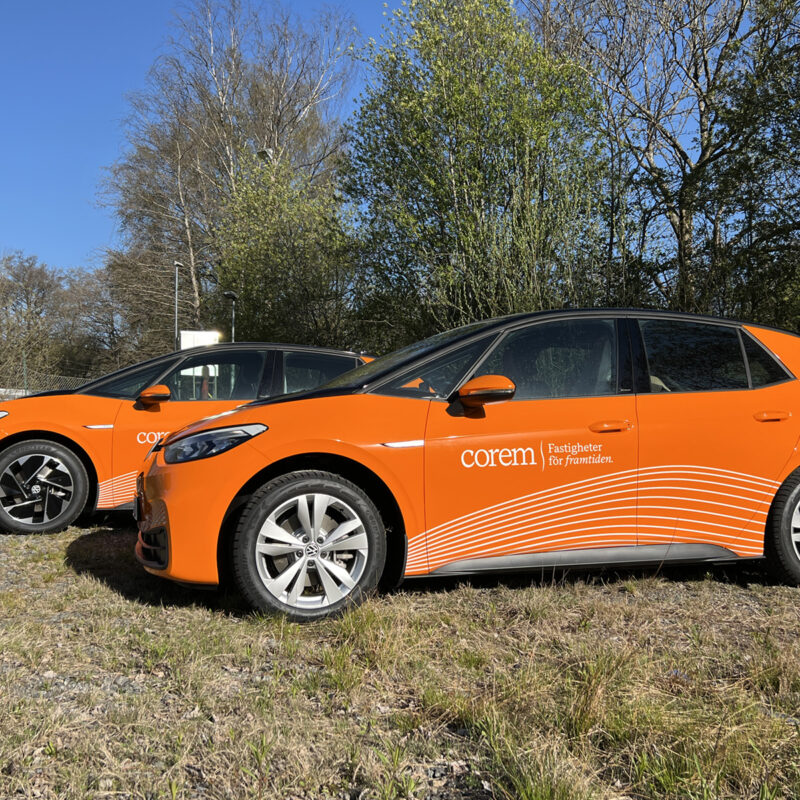 Två bilar står uppställda på en äng. Båda är helfolierade med en orange printad dekor med Corem fastigheter logotype på sidan.