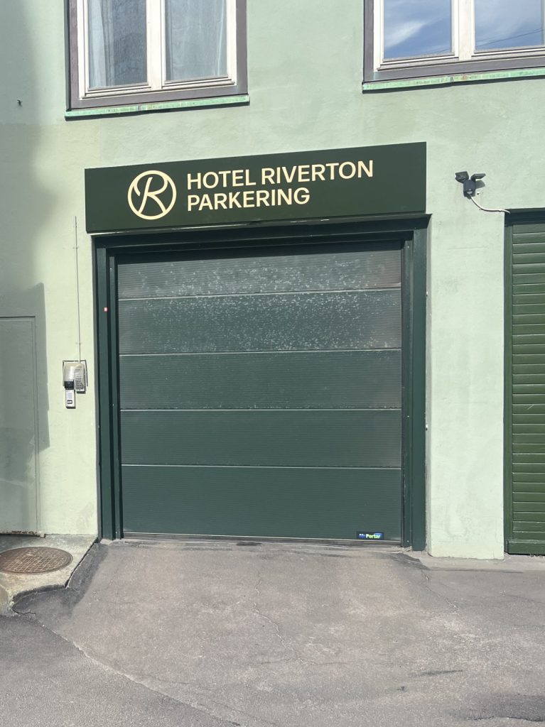 En skylt behöver inte bara visa vägen, den kan också bli ett komplement till en byggnad, en entré eller som här – Fasadskyltar för Hotel Riverton parkering. Infarten till parkeringen för Hotel Riverton pryds nu av en vacker mörkgrön fasadskylt med guldfärgad text i figurskuren aluminium.