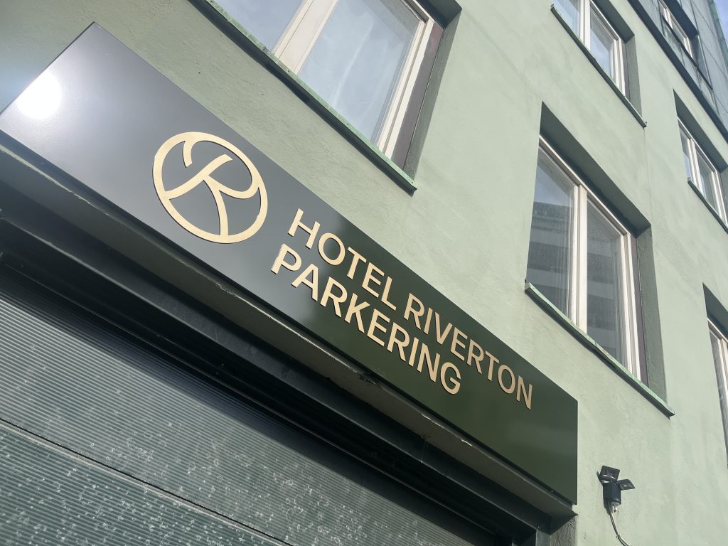 En skylt behöver inte bara visa vägen, den kan också bli ett komplement till en byggnad, en entré eller som här – Fasadskyltar för Hotel Riverton parkering. Infarten till parkeringen för Hotel Riverton pryds nu av en vacker mörkgrön fasadskylt med guldfärgad text i figurskuren aluminium.