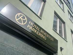 En skylt behöver inte bara visa vägen, den kan också bli ett komplement till en byggnad, en entré eller som här – Fasadskyltar för Hotel Riverton parkering. Infarten till parkeringen för Hotel Riverton pryds nu av en vacker mörkgrön fasadskylt med guldfärgad text i figurskuren aluminium.