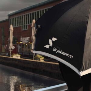 Paraply med tryckt logotype och reflexband. Det kantas av ett reflekterande band runt panelerna. När det är mörkt ute och dålig sikt i regnet tillför det reflekterande bandet runt ytterkanten lite extra synbarhet.