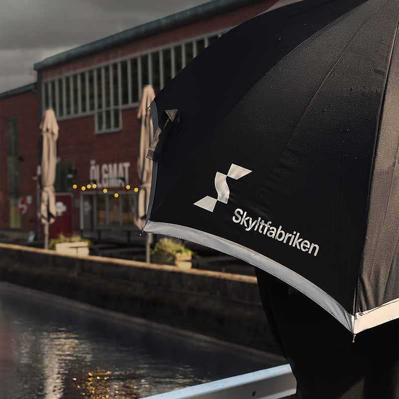 Paraply med tryckt logotype och reflexband. Det kantas av ett reflekterande band runt panelerna. När det är mörkt ute och dålig sikt i regnet tillför det reflekterande bandet runt ytterkanten lite extra synbarhet.