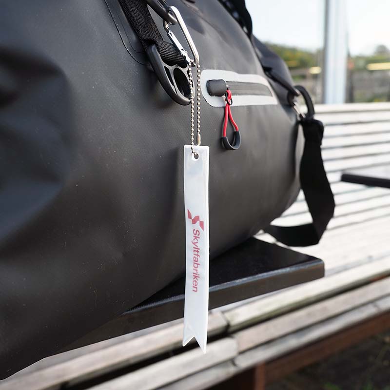 Sätt reflexen på väskan istället för jackan. Ny design i mjuk plast med karbinhake. Tryck kan fås i en eller flera färger.